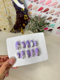 Set of purple nail art designs on a white card held by a hand, with a colorful background.